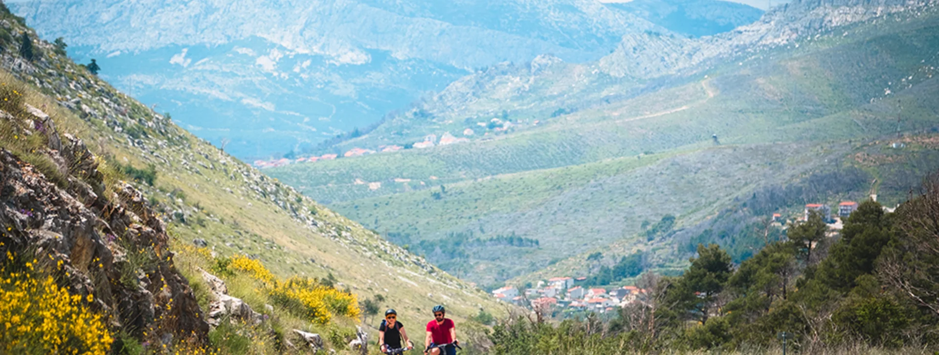 Two cyclists ride along a winding road, surrounded by lush hills and mountains under a partly cloudy sky.