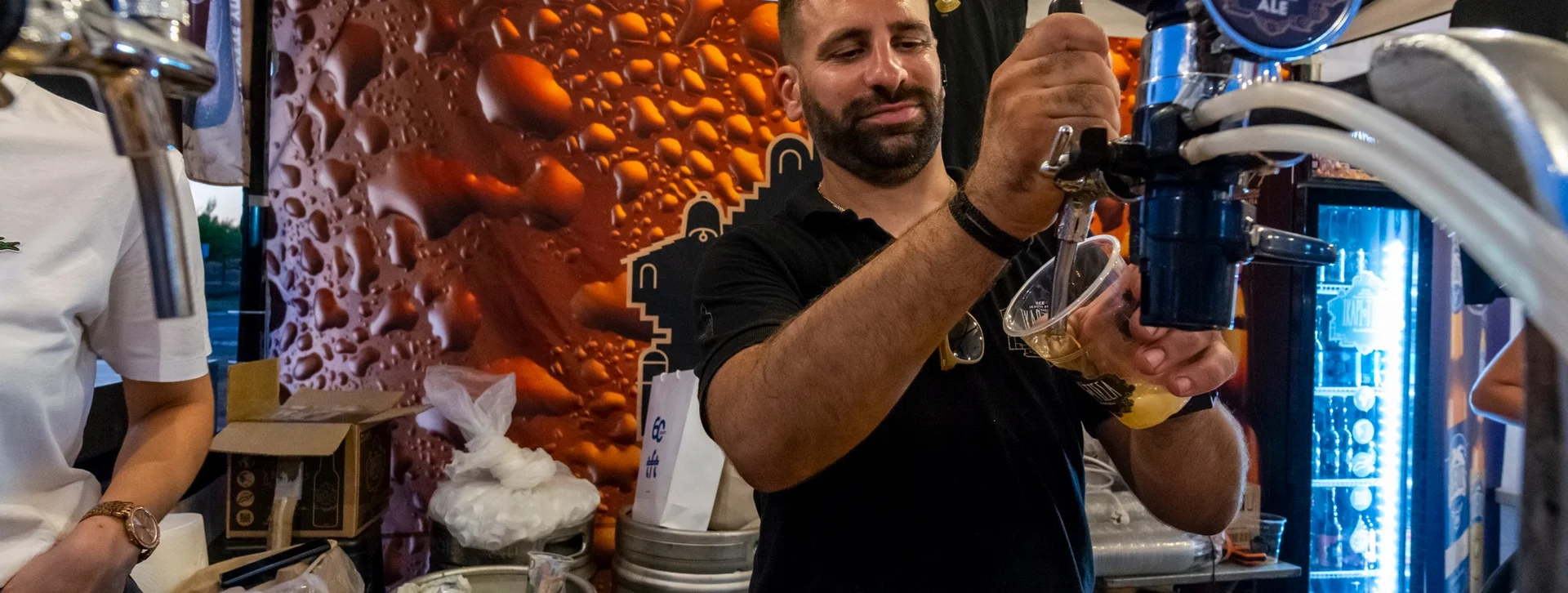 A bartender fills a clear cup with beer from a tap at a lively outdoor market, surrounded by kegs and a decorative backdrop.