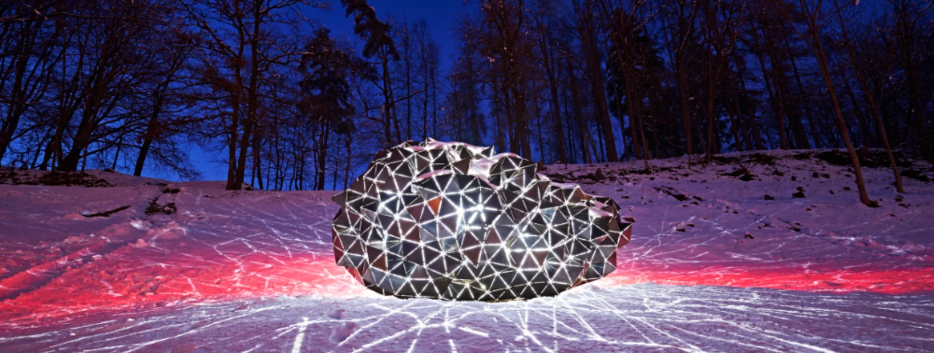 A geometric, illuminated sculpture glows in the snowy landscape, casting intricate light patterns against a twilight sky.