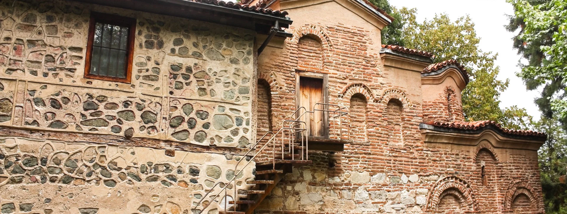 Stone and red-brick façade of Boyana Church, surrounded by lush greenery and featuring a small apse and tower.