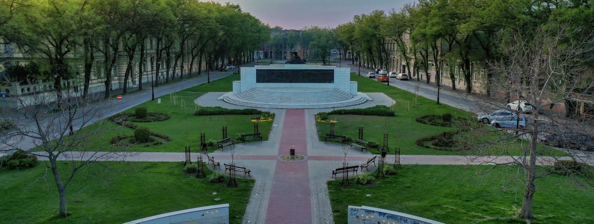 Aerial view of a park featuring a circular plaza with steps, benches, and lush trees under a pastel sky at sunset.