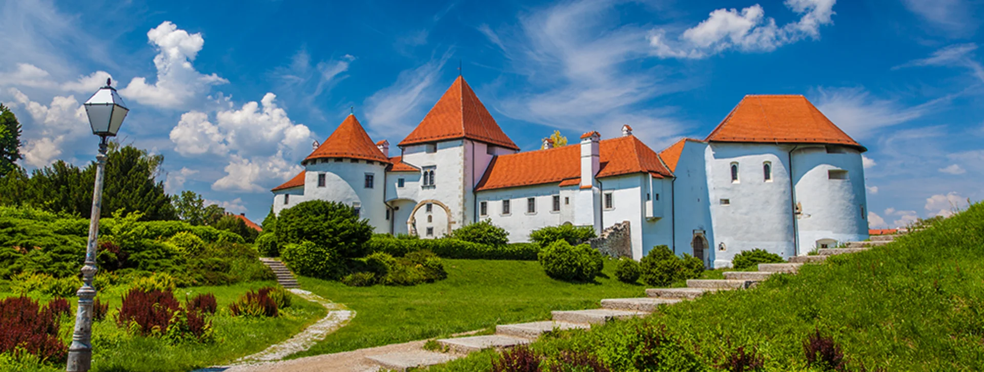 Historic white castle with red roofs, surrounded by lush greenery and a clear blue sky, set against vibrant clouds.