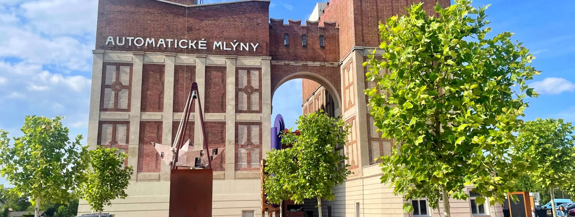 Historic Automatic Mills building with an archway, flanked by green trees and a modern sculpture in a sunny outdoor setting.