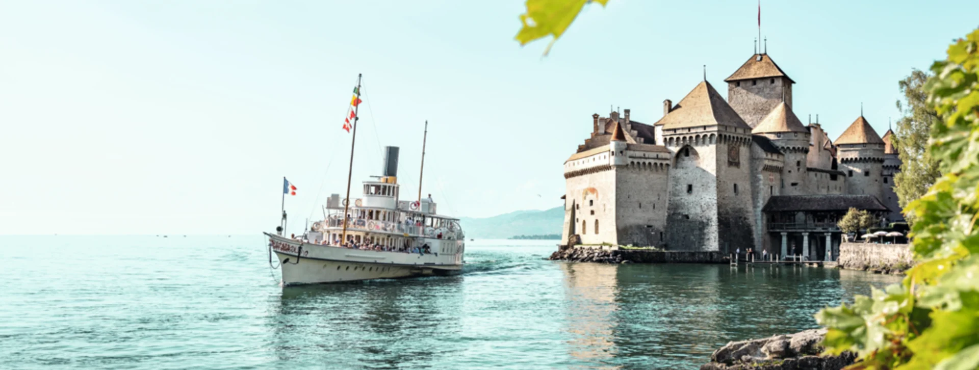 A vintage paddle steamer glides across a serene lake, past a medieval castle with turrets and a blue sky in the background.