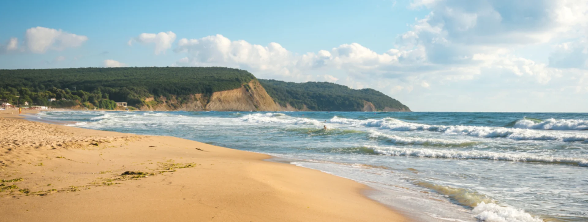Sandy beach with waves and distant forested cliffs.