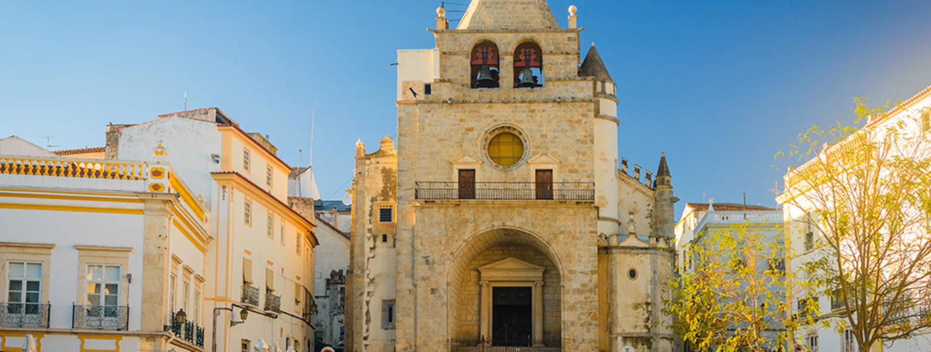 Sunny view of Praça da República in Elvas, Portugal, showing the cathedral with its stone facade and white buildings beside it, all bathed in bright daylight.