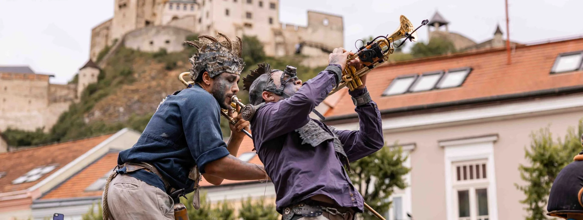 Deux artistes de rue en costumes steampunk jouent des instruments à vent modifiés, avec le château de Trenčín sur la colline derrière eux.