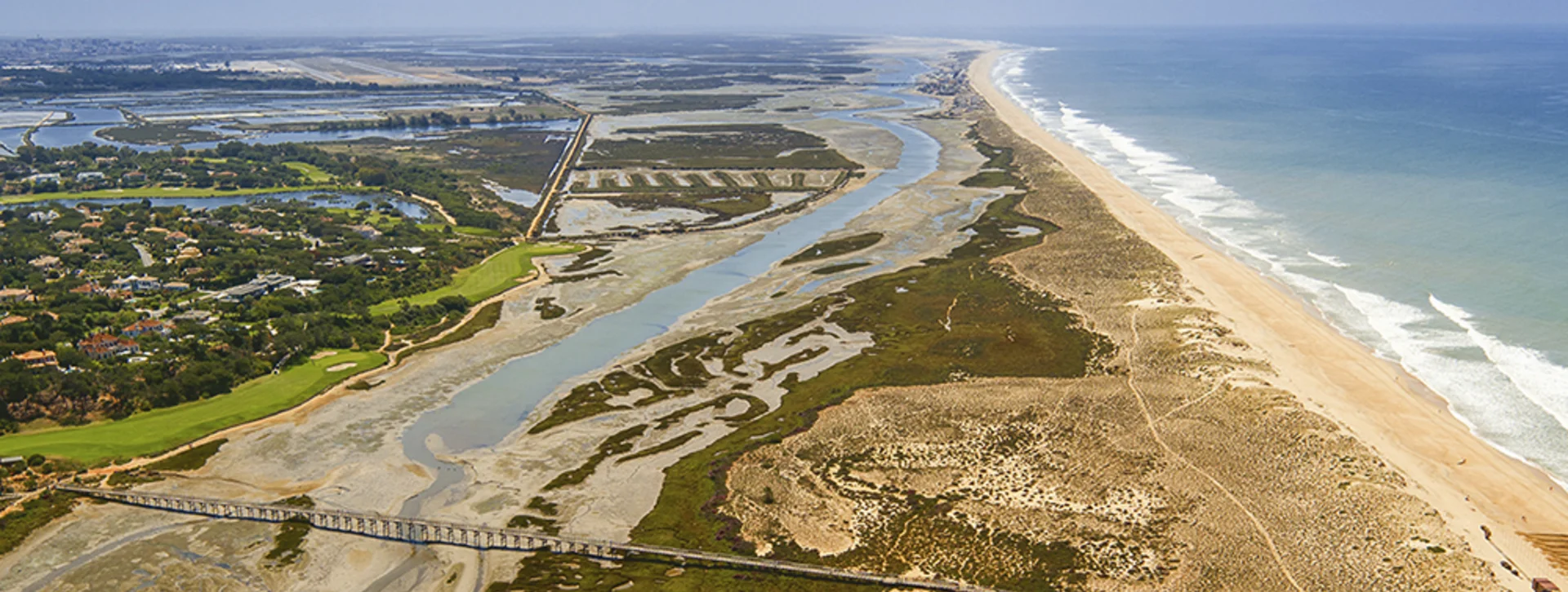 アルガルヴェ地方のキンタ・ド・ラーゴ・リゾートとリア・フォルモーザ沿岸潟の空撮。砂丘、塩性湿地の水路、木製の遊歩道橋が見えます。