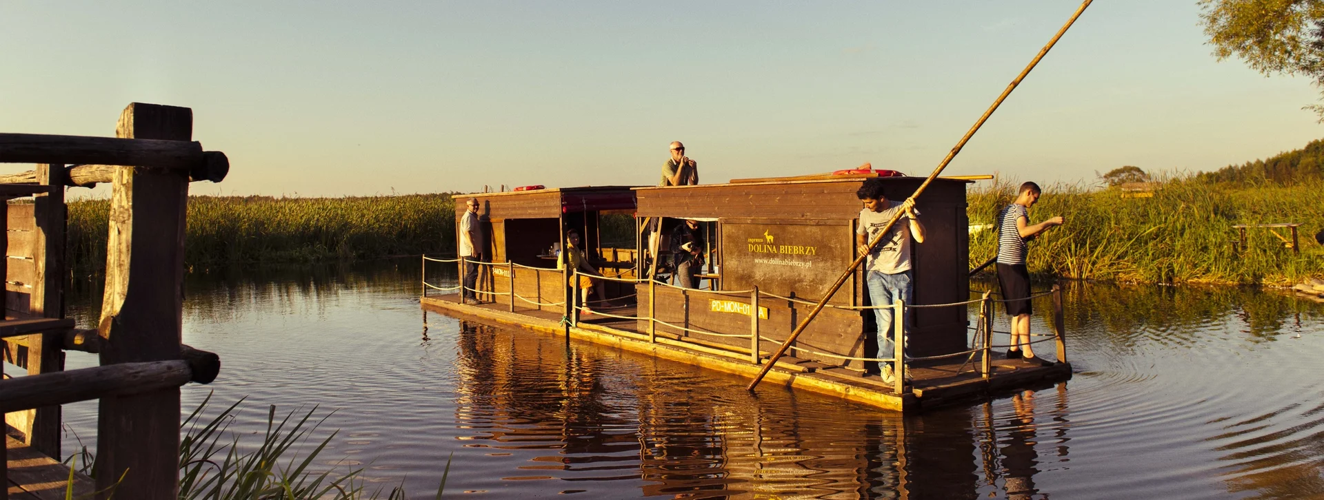 People on a wooden raft navigating a calm river at sunset.