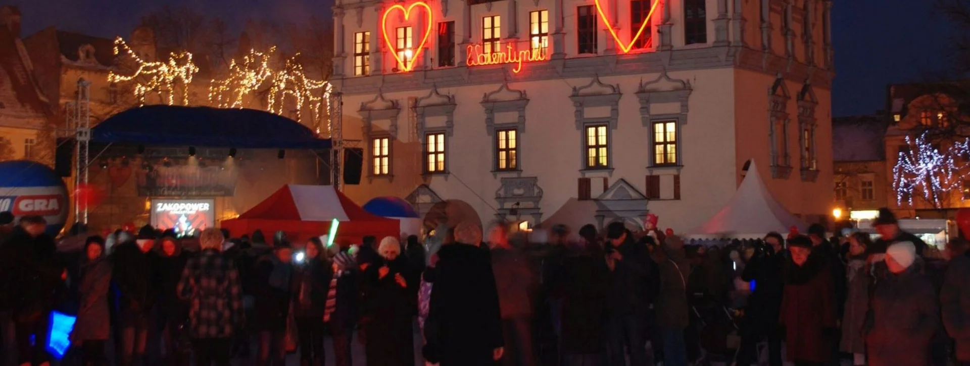 Crowd gathered in a historic town square with heart-shaped lights at night.