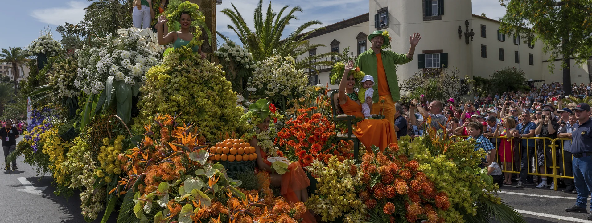 Colorful parade float with large flower arrangements and people waving, crowd watching in background.