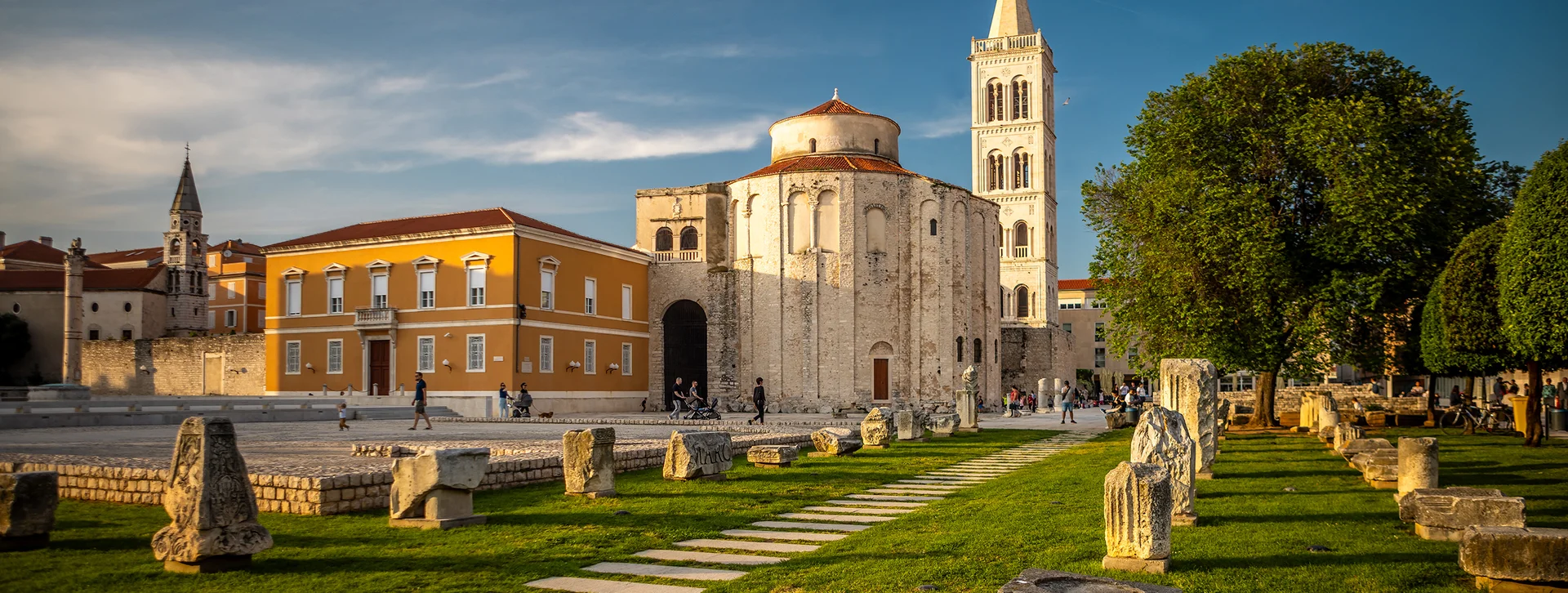 Historic church with a tall bell tower, stone ruins, and green lawn under a blue sky.