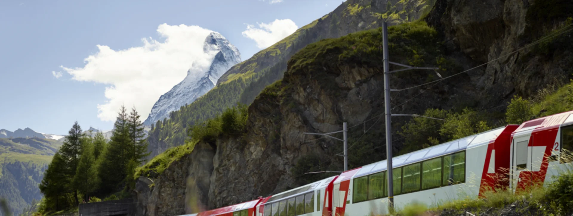 Red and white train passing through a mountainous landscape with wildflowers and blue sky.