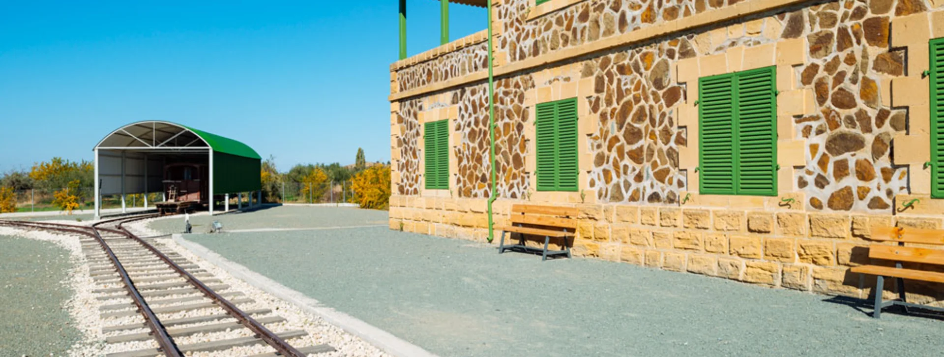 Stone building with green shutters beside empty railway tracks and benches under a clear blue sky.