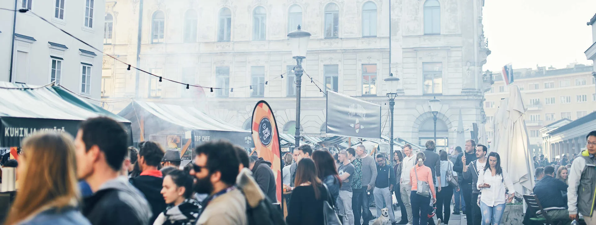 Crowded outdoor market with people, stalls, and a historic building in the background.