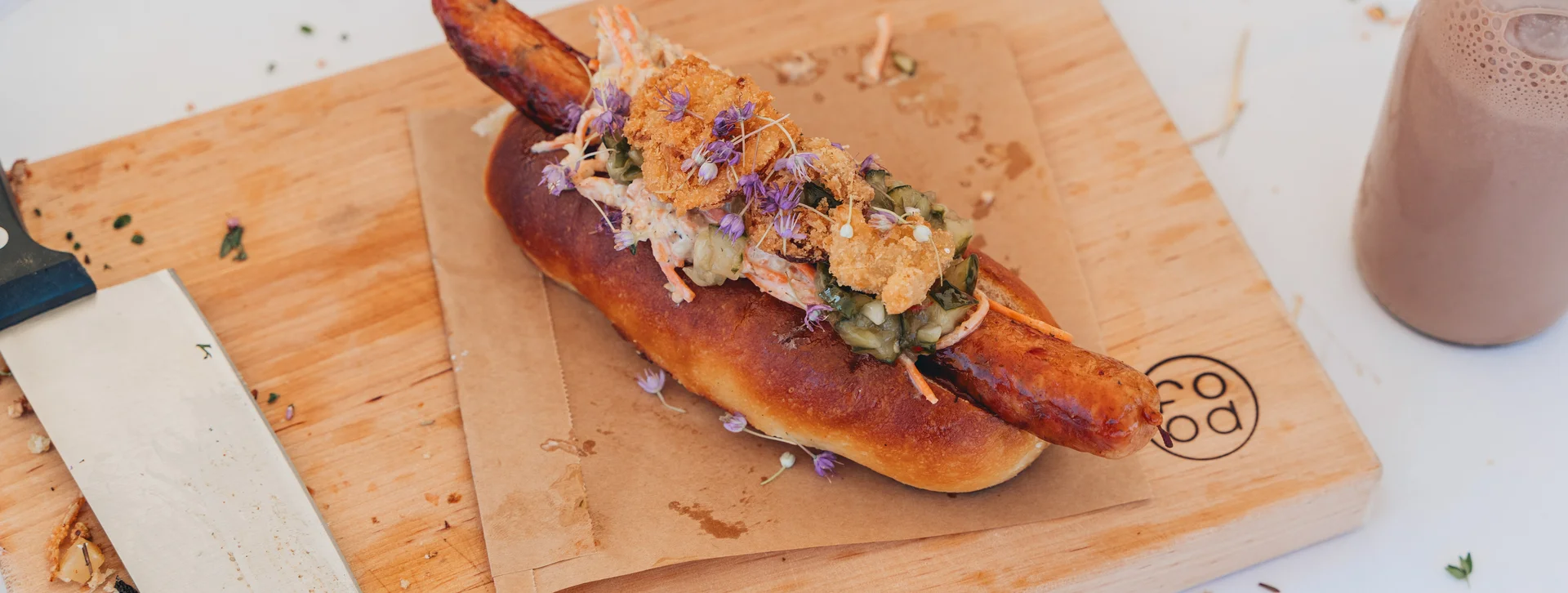Gourmet sausage in a bun topped with slaw on a wooden board, knife beside it.