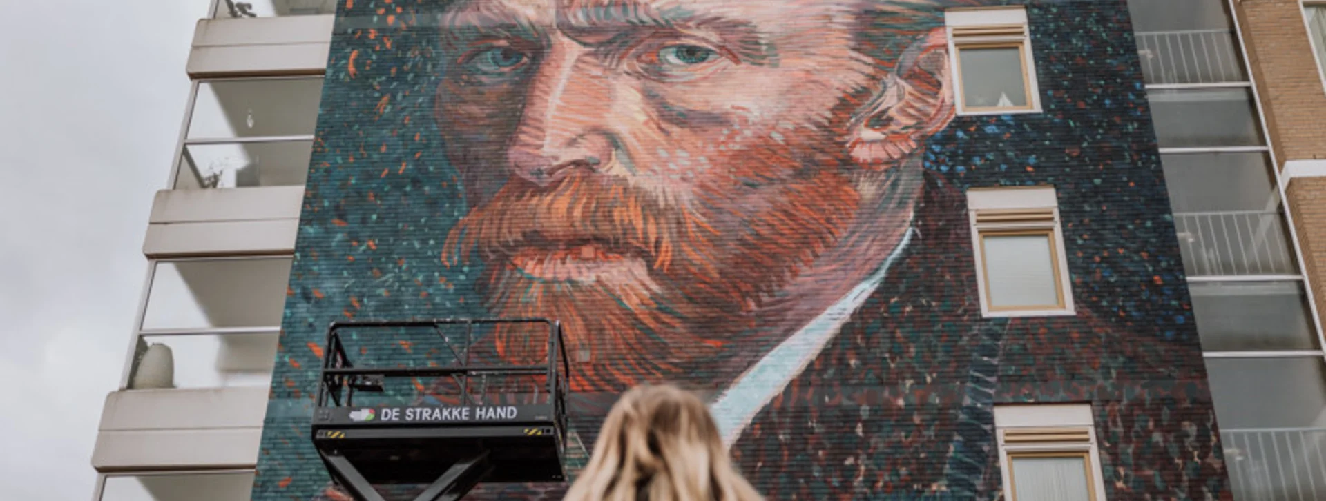 Person looking up at a large mural of Vincent van Gogh on a building wall.