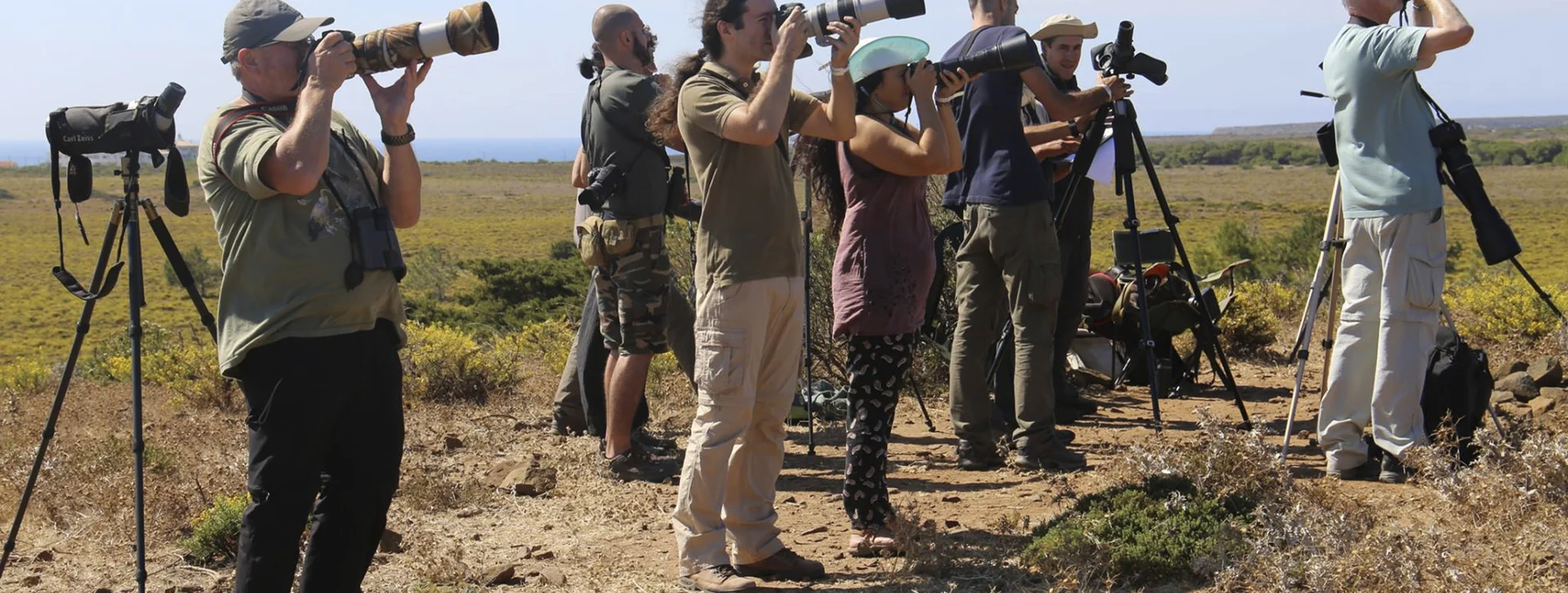 Group of people birdwatching with cameras and binoculars in a grassy field.