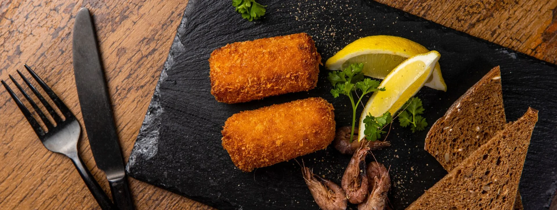 Two breaded croquettes, brown bread slices, lemon wedges, and parsley on a black slate plate.