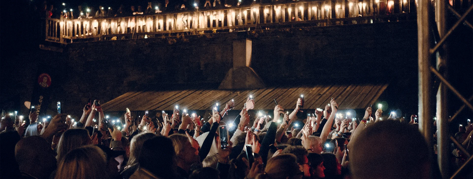 Crowd at night holding up lit phones, with bridge lights shining above.