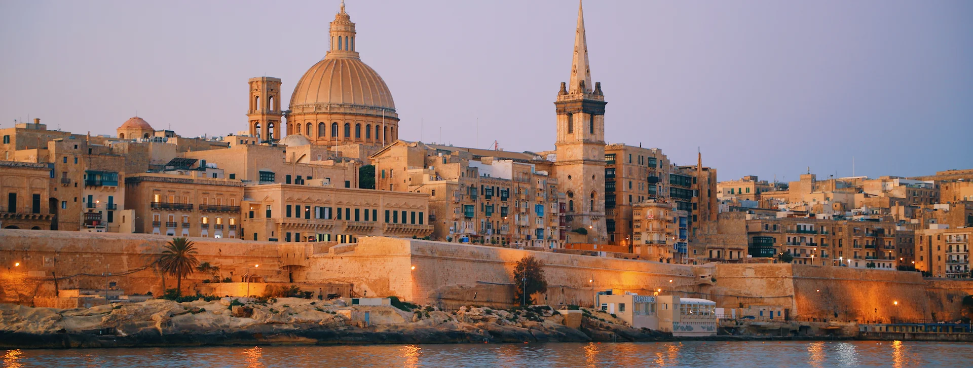 Historic stone buildings and domed church along a waterfront at sunset.