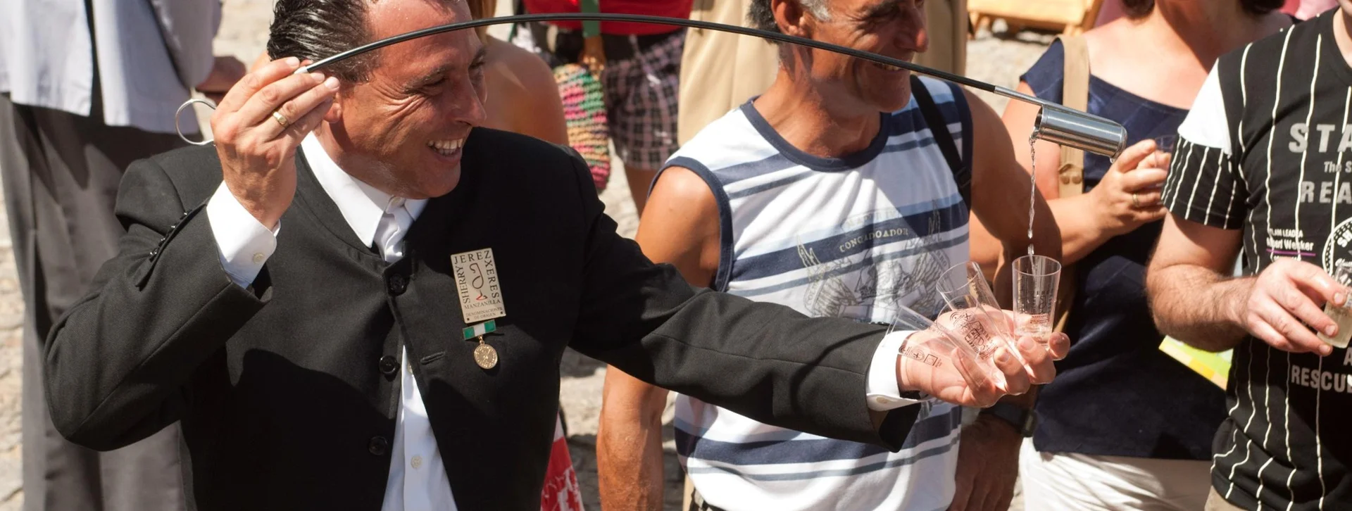 Man in formal attire pours wine with a long spout, smiling crowds watching outdoors.