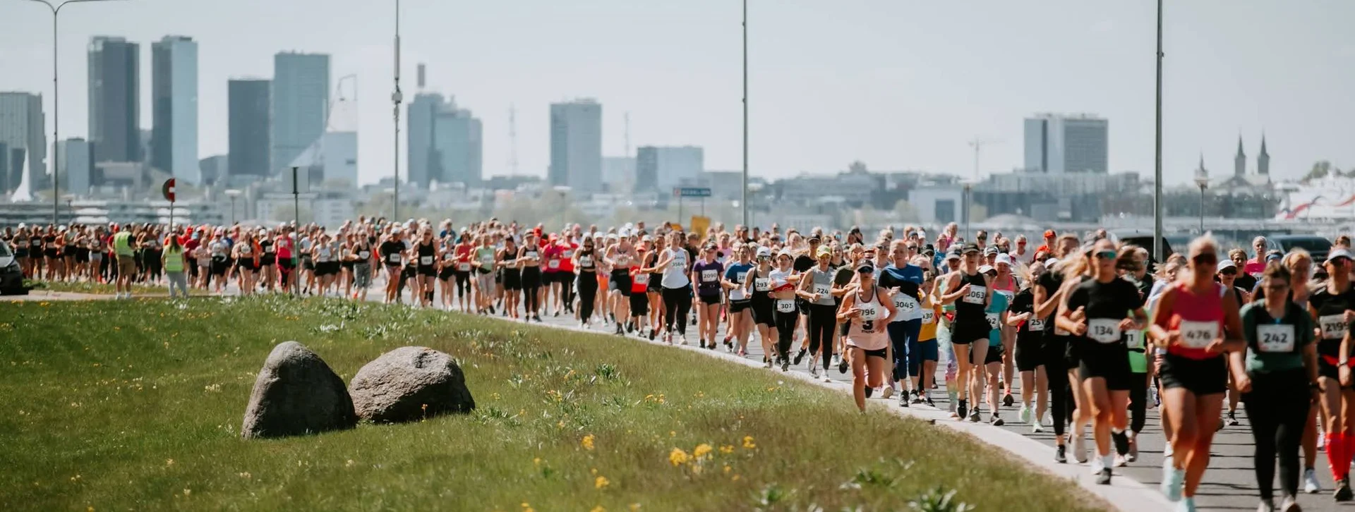 A large group of runners on a road by grassy fields, city skyline in the background.