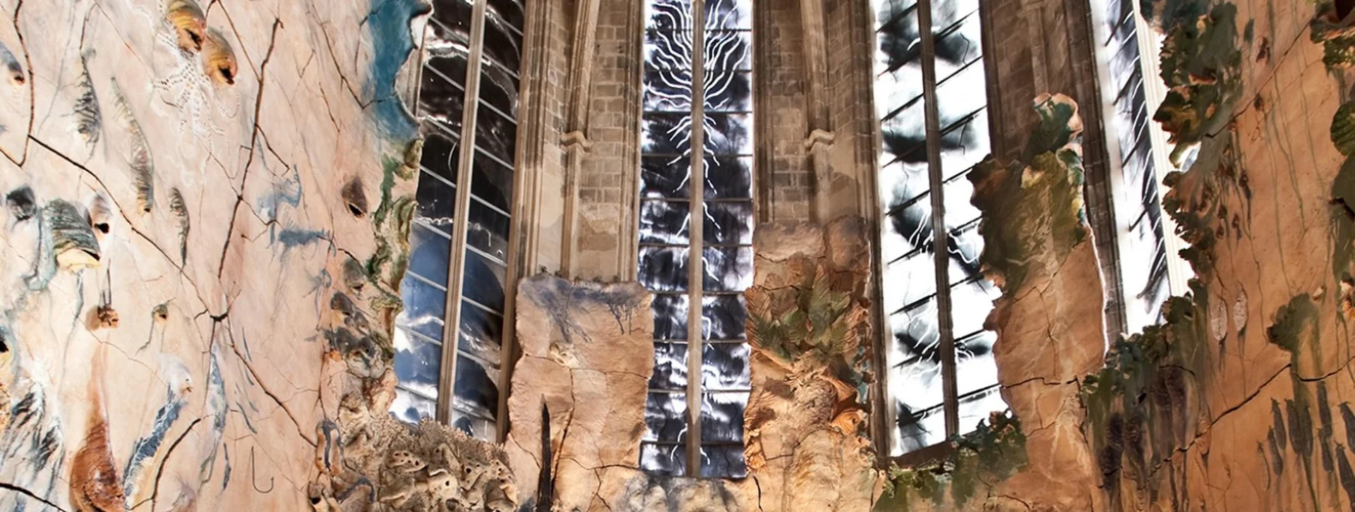 Tall stained glass windows and painted walls inside a cathedral with tall arches and plants.