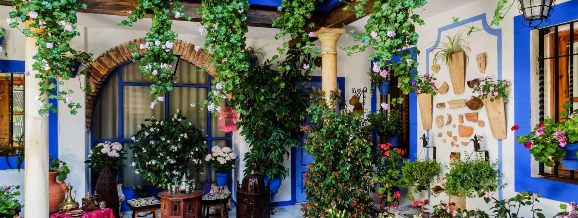 Colorful courtyard with lush hanging plants, flowers, columns, and blue-trimmed windows.