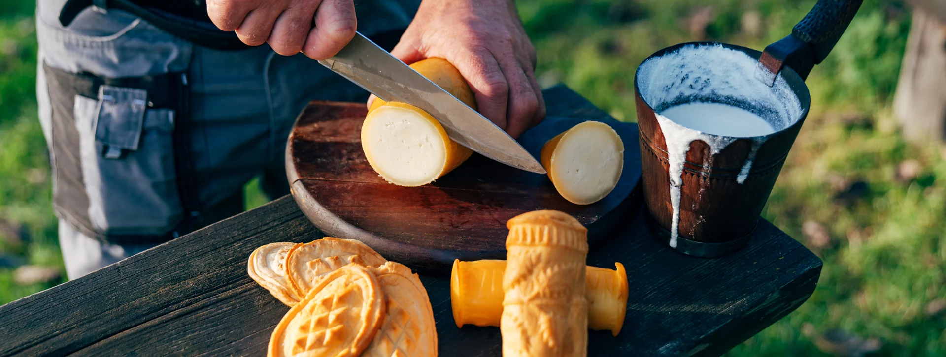 Person slicing smoked cheese on a wooden board outdoors, cup of milk nearby.