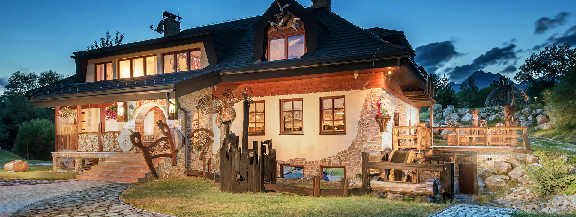 Cozy wooden house with lit windows at dusk, stone pathway, and rustic decorations.