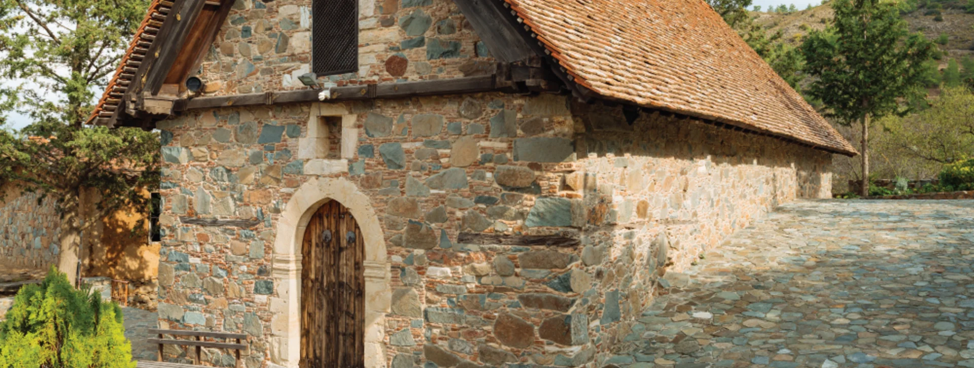 Old stone building with a tile roof, surrounded by trees and a stone courtyard.