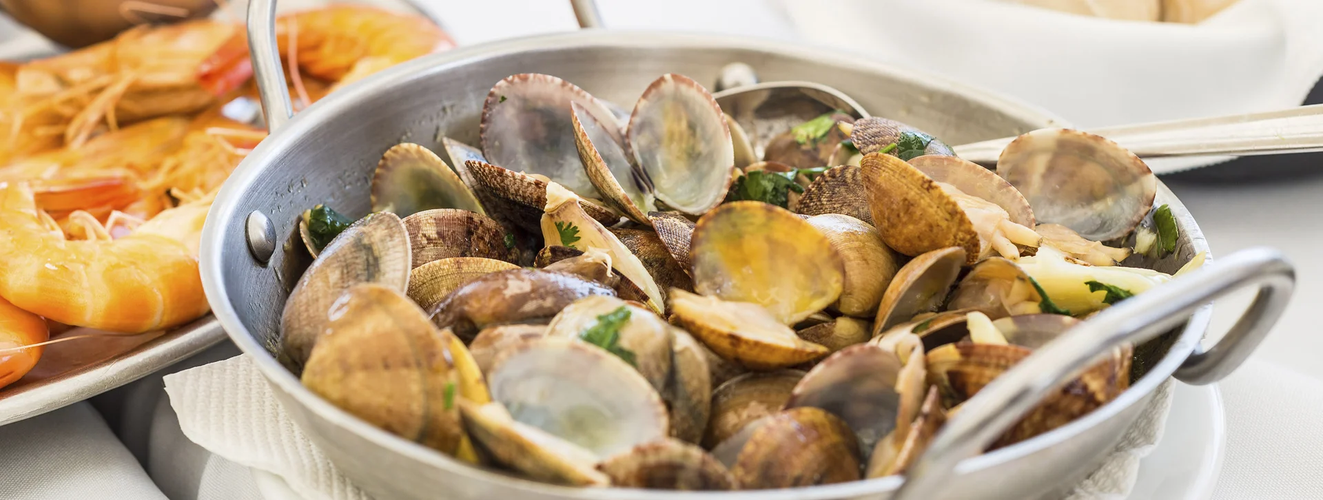 Steamed clams in a metal pan garnished with herbs on a white tablecloth.