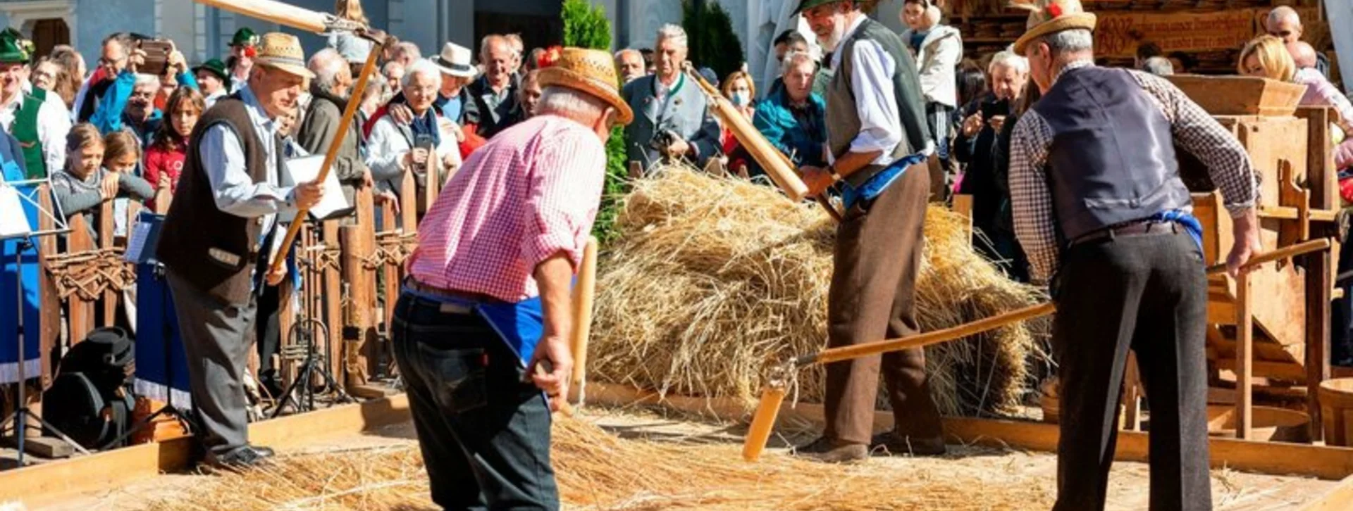 Men in traditional clothes threshing grain on a wooden platform at a festival.