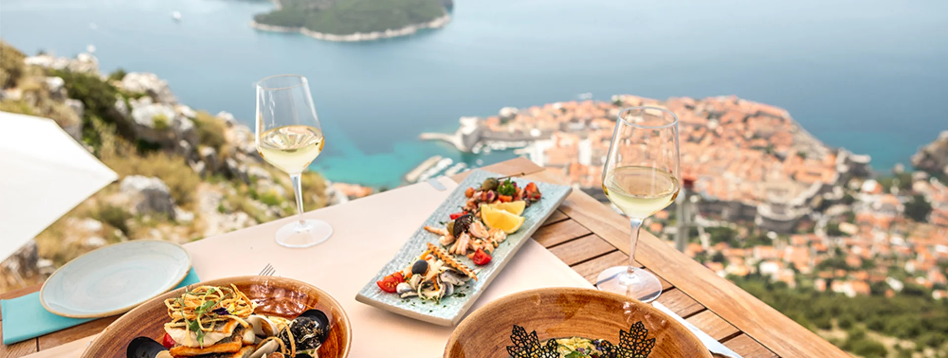 Seafood dishes and wine on a table overlooking a coastal city and blue sea.