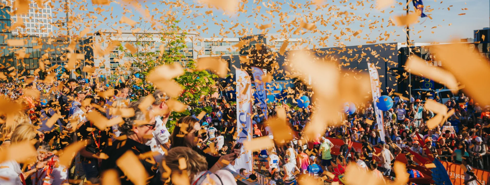 Orange confetti falling over a large, festive crowd outdoors under blue sky.