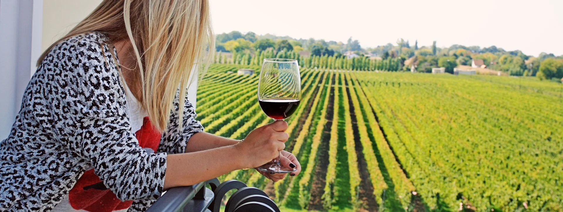 Woman holding a glass of red wine overlooking a green vineyard from a balcony.