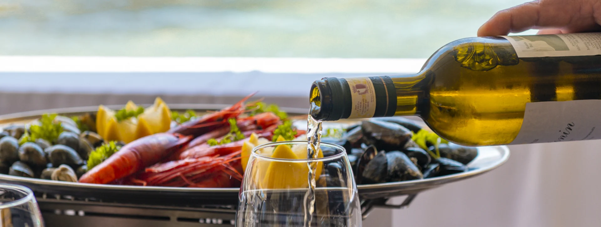 White wine being poured into a glass next to seafood by the seaside.