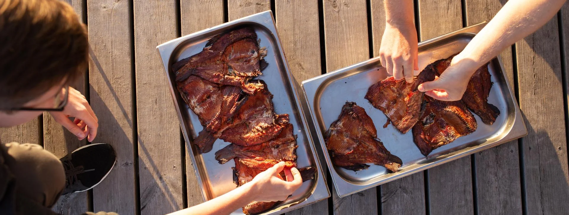Two people picking ribs from trays on a wooden deck in sunlight.