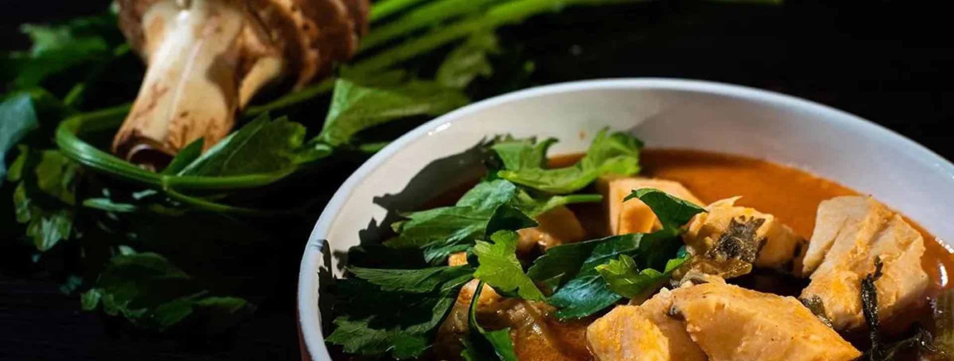Bowl of curry with taro chunks, garnished with fresh herbs, next to raw taro and greens.
