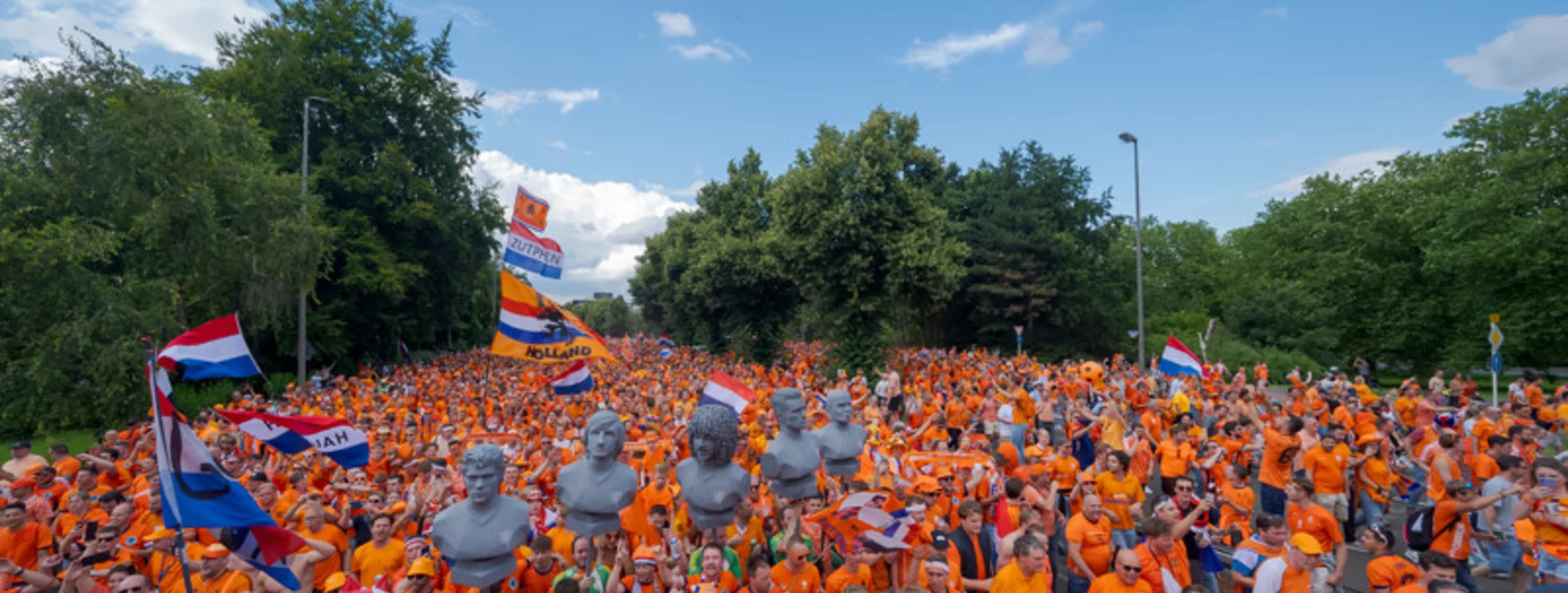 Large crowd in orange shirts with flags gathered outdoors under a blue sky.