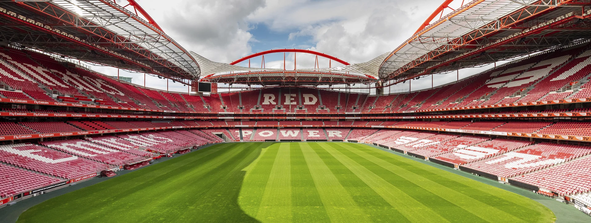 Empty soccer stadium with red seats and a green field under a partly cloudy sky.