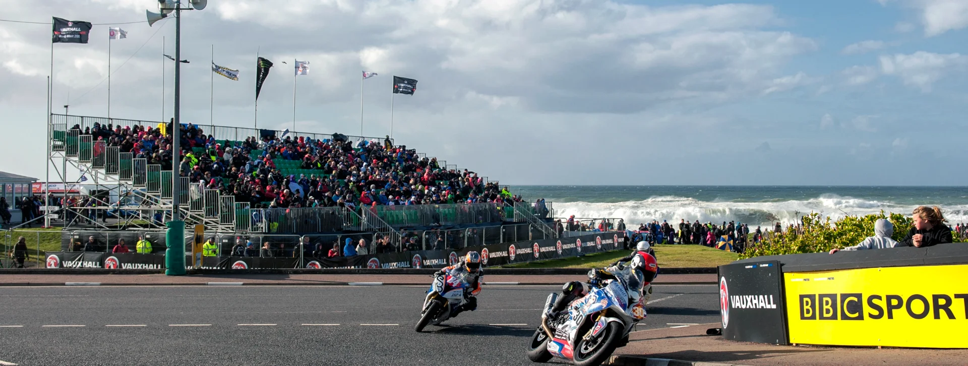 Two motorcyclists race on a track by the ocean with a crowd in the stands.