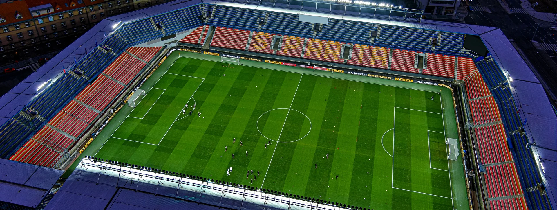 Empty football stadium with green field and red seating, lit up at dusk.