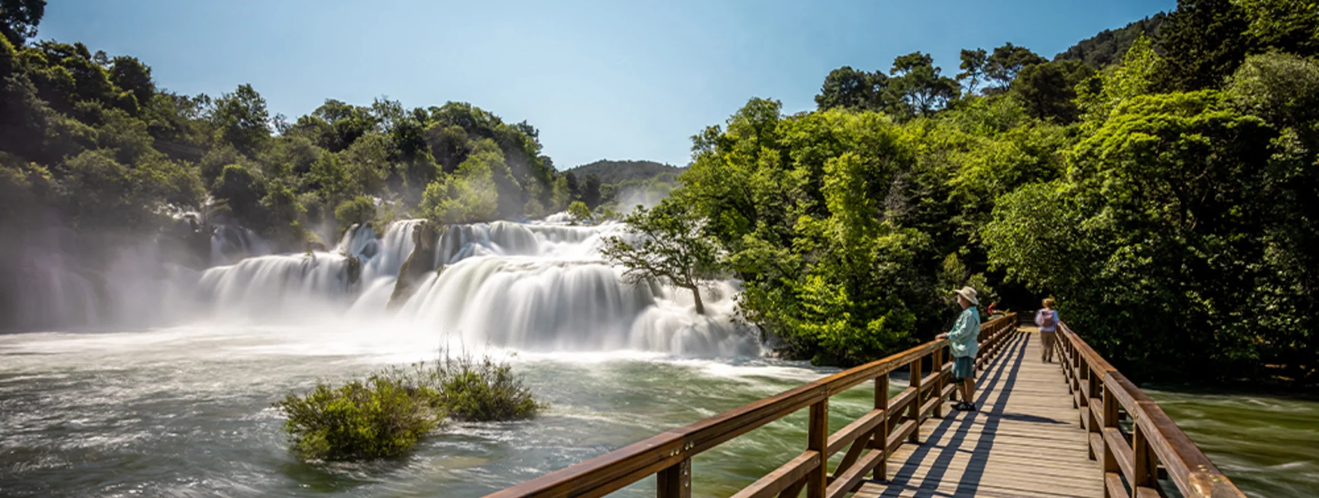 Wooden footbridge leads to a large waterfall surrounded by lush green trees on a sunny day.