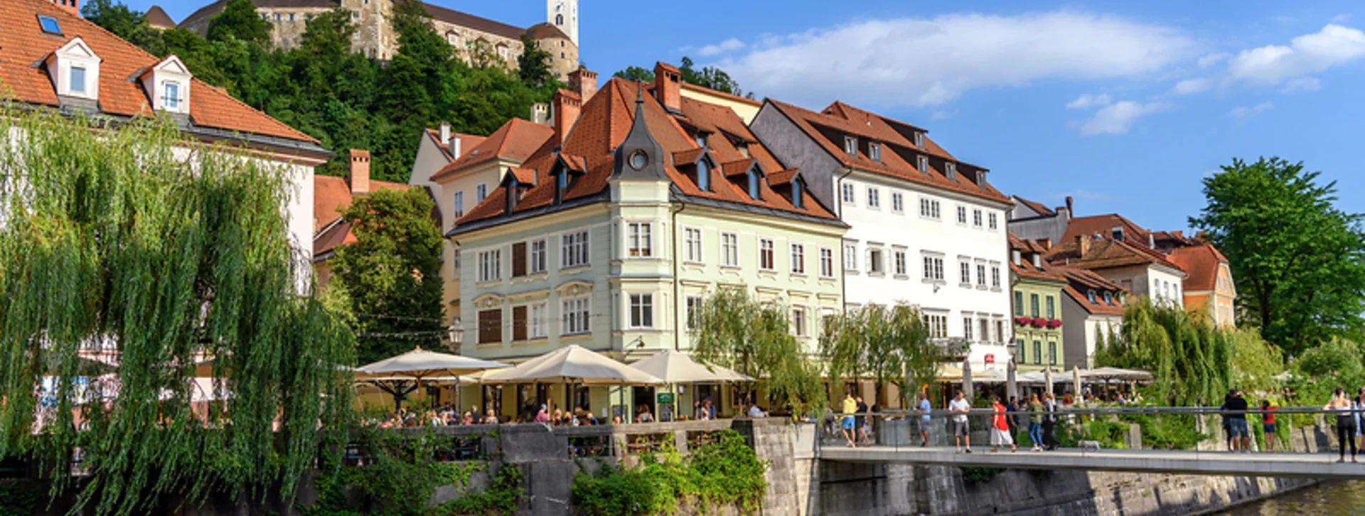 Colorful riverside buildings with outdoor cafes, lush greenery, and a castle on a hilltop.