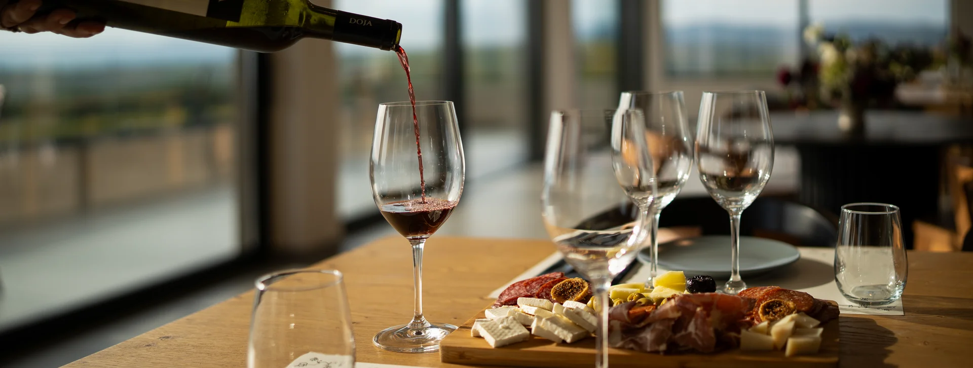 Red wine being poured into a glass beside a cheese and charcuterie board on a dining table.