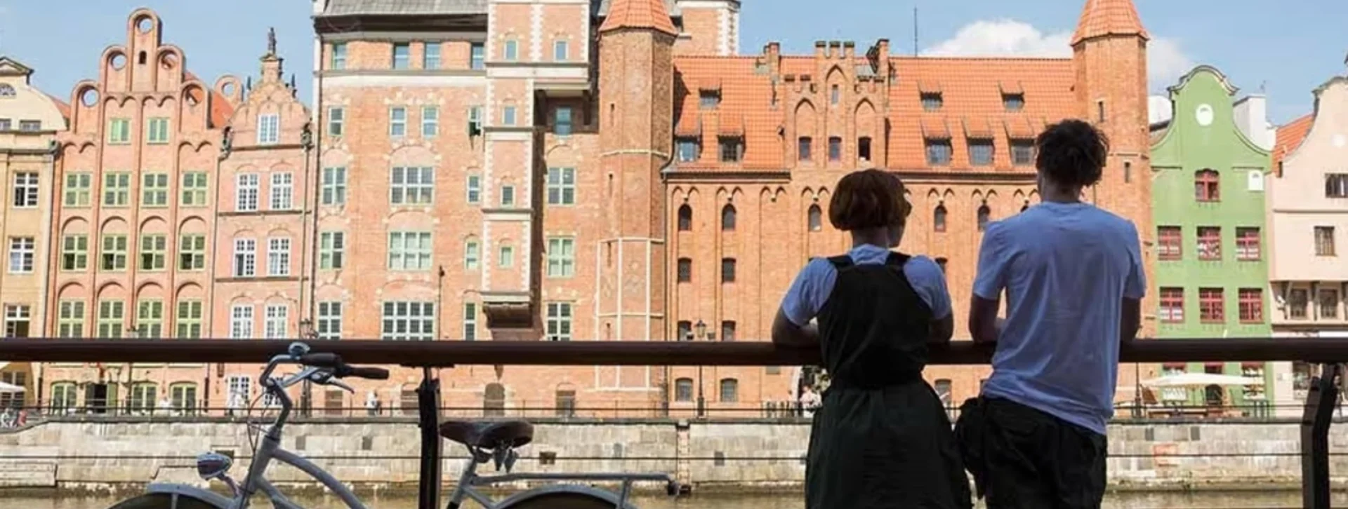 Two people and a bicycle by a river, colorful historic buildings in the background.