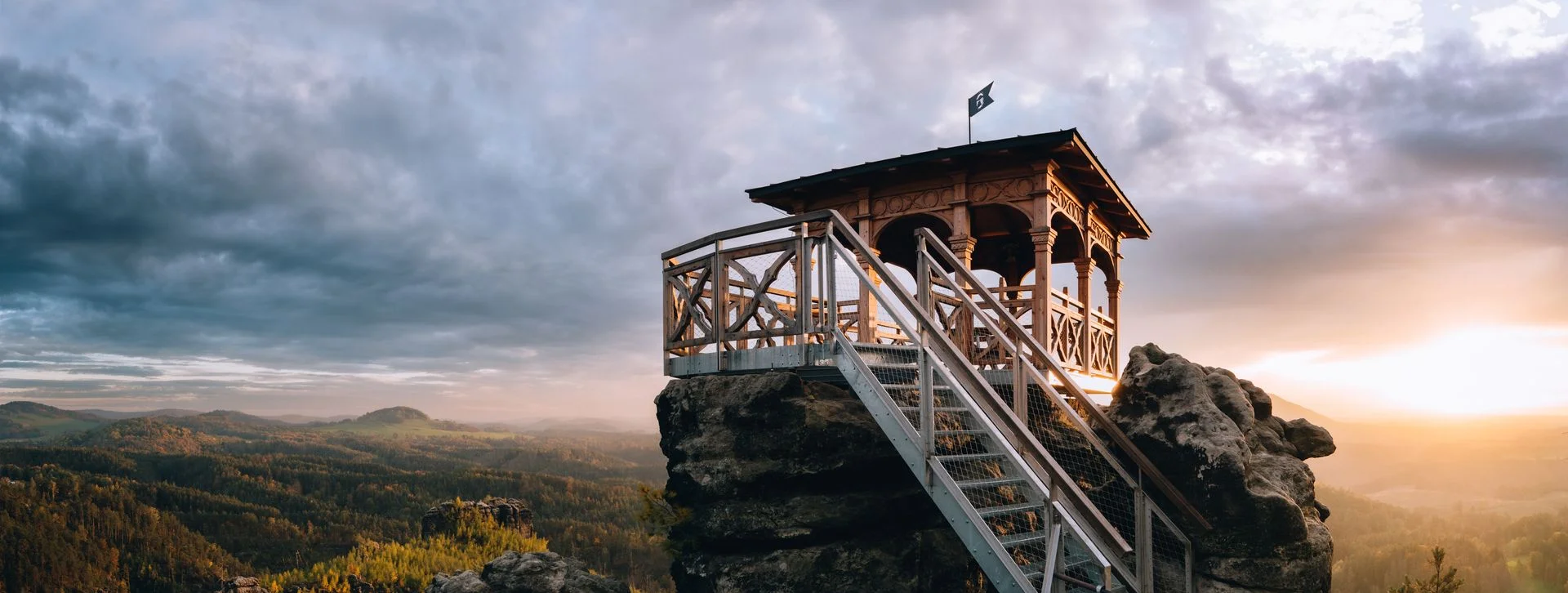Wooden lookout tower on rocky cliff at sunrise overlooking a forested valley.