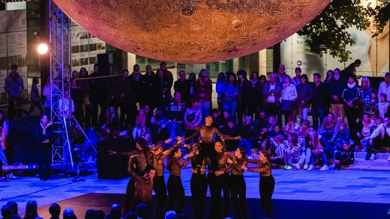 Performers under a large, textured disc with an audience watching at night.